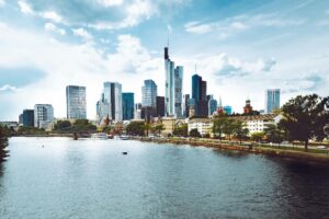A modern cityscape of Frankfurt with skyscrapers reflecting in the river under a blue sky.